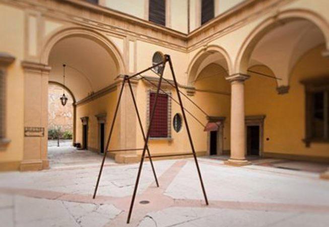 A modern sculpture consisting of an old iron swing with the seat still suspended in the air, installed in the middle of an ancient courtyard. Sculpture by Dado
