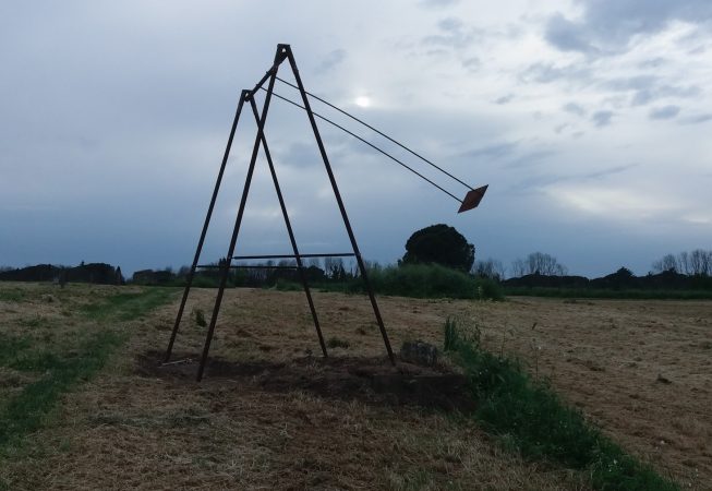 A modern sculpture installation consisting of an old iron swing with the seat still suspended in the air in the middle of a grass field. Sculpture by Dado