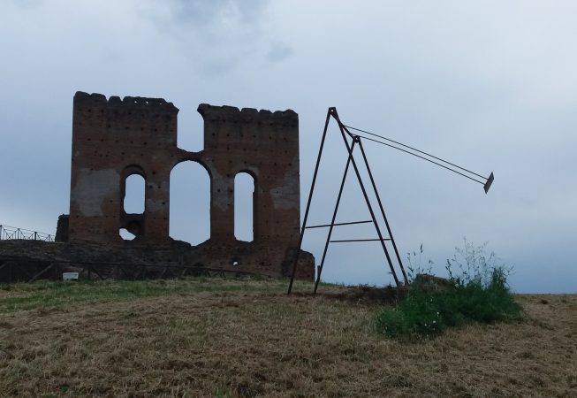 A modern sculpture installation consisting of an old iron swing with the seat still suspended in the air with an ancient roman wall ruin in the background. Sculpture by Dado