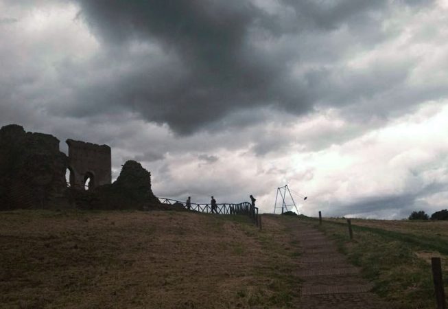 Wide view of an hill, on an overcast day, on top of which are located an old roman ruin and a modern art installation consisting of an old iron swing with the seat still suspended in the air. Installation by Dado