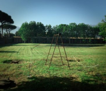 A modern sculpture installation consisting of an old iron swing with the seat still suspended in the air, in the middle of a grass field. Sculpture by Dado