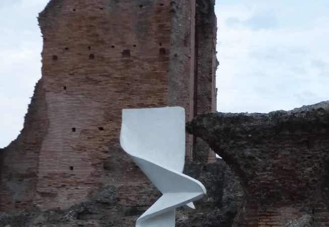 Portrait view of a modern art installation, placed in front of old roman ruins, consisting of a white concrete column in the form of a vertical ribbon with a sharp fold in the middle producing some interesting shades contrast. Installation by Dado