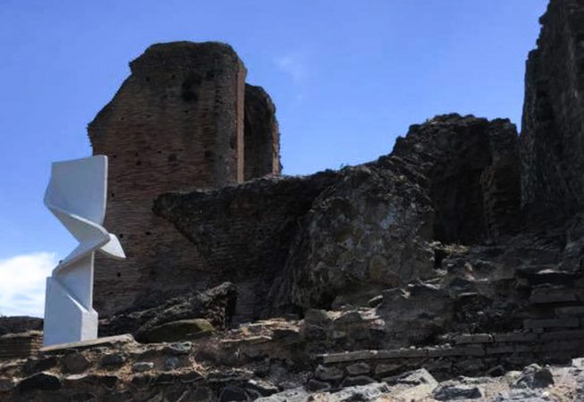 Wide view of a modern art installation, placed in front of old roman ruins, consisting of a white concrete column in the form of a vertical ribbon with a sharp fold in the middle producing some interesting shades contrast. Installation by Dado