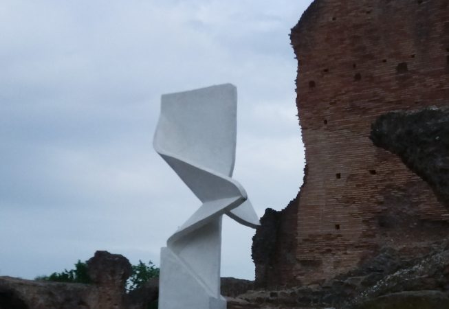 Portrait view of a modern art installation, placed in front of old roman ruins, consisting of a white concrete column in the form of a vertical ribbon with a sharp fold in the middle producing some interesting shades contrast. Installation by Dado