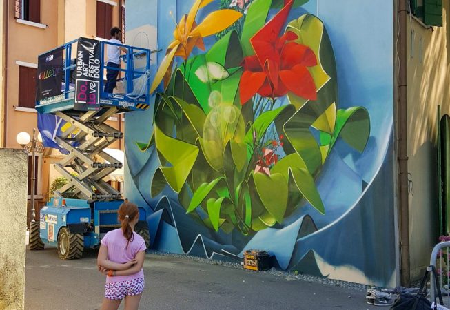 Dado painting a Graffiti-mural spherical lettering with multi colored flowers-ground view of the entire wall and the artist on an industrial elevator while a kid in pink dress is watching