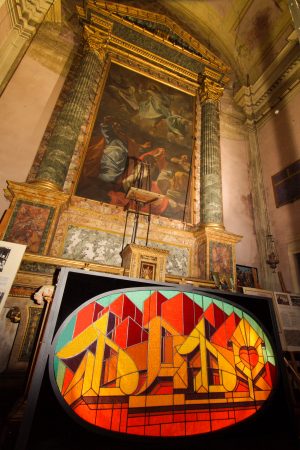 Inside an old church is placed for display a stained glass window made colored glass pieces representing the word Dado in a graffiti style at the bottom of stylized skyscrapers. bottom left view