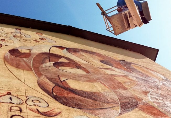 A wall with a mural painting depicting spherical wildstyle graffiti forming the tag Dado with the artist working on the wall on a lift basket. Bottom up view