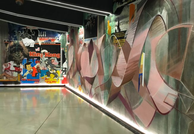 Right total view of a decorated garage wall recreating a street underground atmosphere with spray can lettering artworks and torn paper posters. Artwork by Dado.