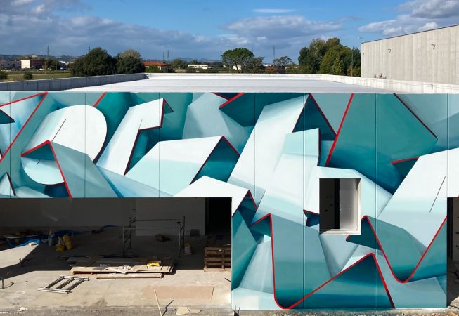 Front view of a graffiti paint work on the external walls of a big building representing flat ribbons bent in geometrical shapes, light blue and white shades with thin red outlines. Artwork by Dado