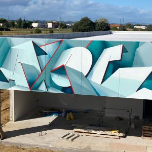 Closer front view of a graffiti paint work on the external walls of a big building representing flat ribbons bent in geometrical shapes, light blue and white shades with thin red outlines. Artwork by Dado