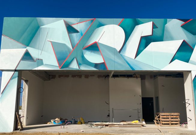 Front ground level view of a graffiti paint work on the external walls of a big building representing flat ribbons bent in geometrical shapes, light blue and white shades with thin red outlines. Artwork by Dado