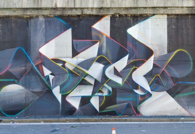 Mural painting representing weaving stiff ribbons forming the name of the artist Dado, in dark grays and white tones with sharp cutting multi color outline. Total view