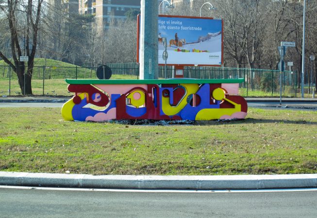 A graffiti style bright colored sculpture in the middle of a traffic circle in a public road path. Installation by Dado