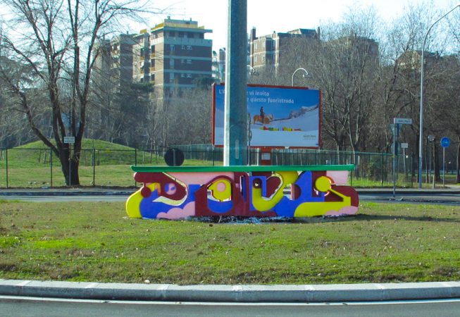 A graffiti style bright colored sculpture in the middle of a traffic circle in a public road path. Installation by Dado