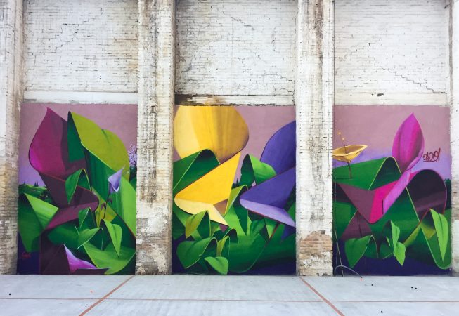 A triptych mural painting of flowers in three different colors on a white bricks wall in a wide open space, total view. Artwork by Dado