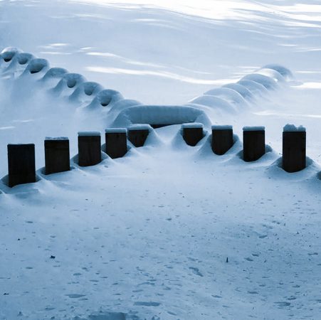 Front view of a wood sculpture composed from a series of tall wooden boards planted in the ground forming a big X just in the middle of a mountain path, covered by the snow.