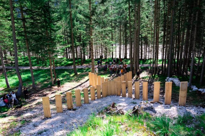 Back view of a wood sculpture composed from a series of tall wooden boards planted in the ground forming a big X just in the middle of a mountain path in a sunny spring day. A group of hikers are approaching the mountain path.