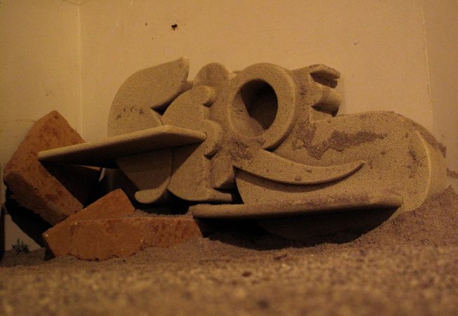 A grey, rock-style, sculpture formed of letters in different styles which composing the name "Sere". The sculpture is laying on the floor along with some thrown brick. Artwork by Dado