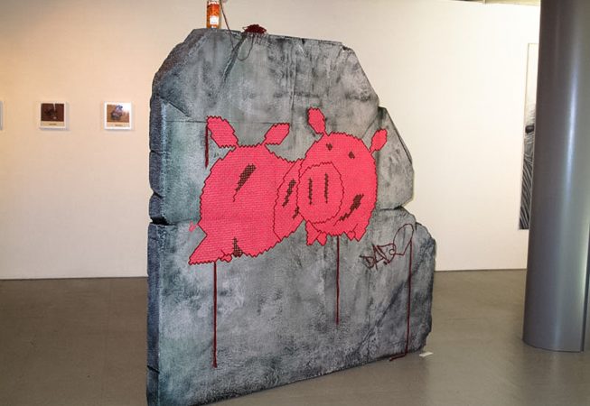 A big rock sculpture with two wool made flat pig characters stitched in the middle. Artwork by Dado