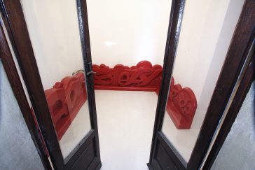 Front view of the installation inside a small room of abas-relief sculpture in the form of a graffiti style baseboard all red color depicting the artists tags Dado and Stefy, decorated with big bubbles in the background