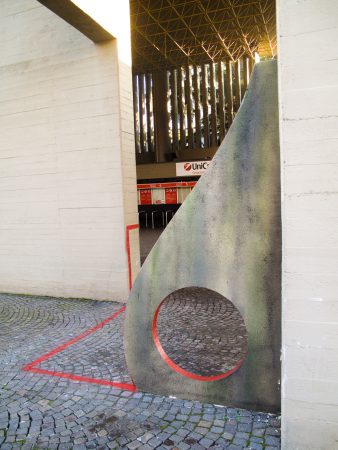Sculpture representing an almost flat "D" sinuous letter installed next to a concrete wall. The red outline of the letter extends from the artwork and runs on the ground. Close view