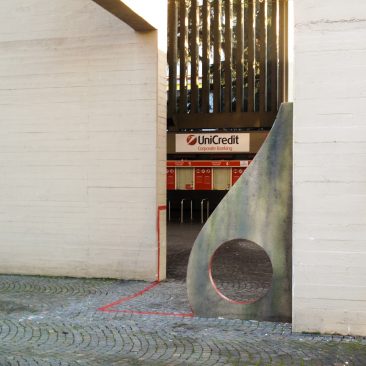 Sculpture representing an almost flat "D" sinuous letter installed next to a concrete wall. The red outline of the letter extends from the artwork and runs on the ground. Left view