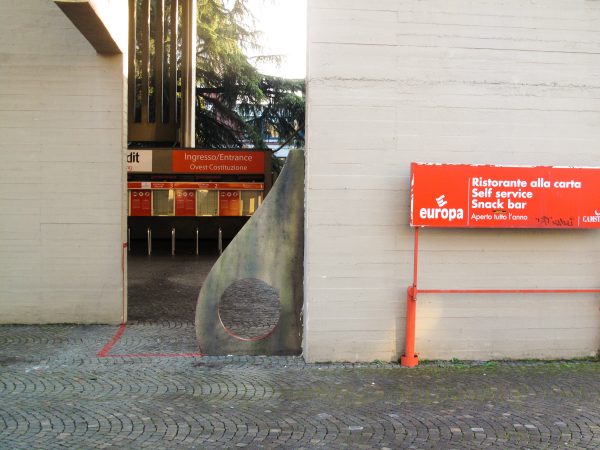 Sculpture representing an almost flat "D" sinuous letter installed next to a concrete wall. Side wide view