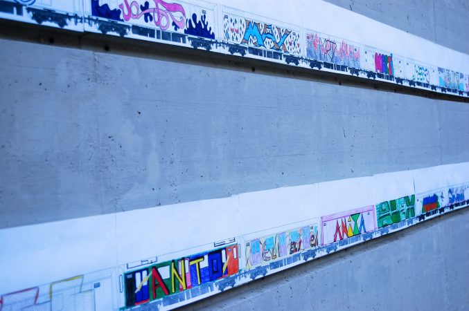 Closeup view of a gray concrete wall with two lines of paper sheets with drawings, each one depicting a train wagon with a graffiti on the side. The sheets are stitched together to form al long train