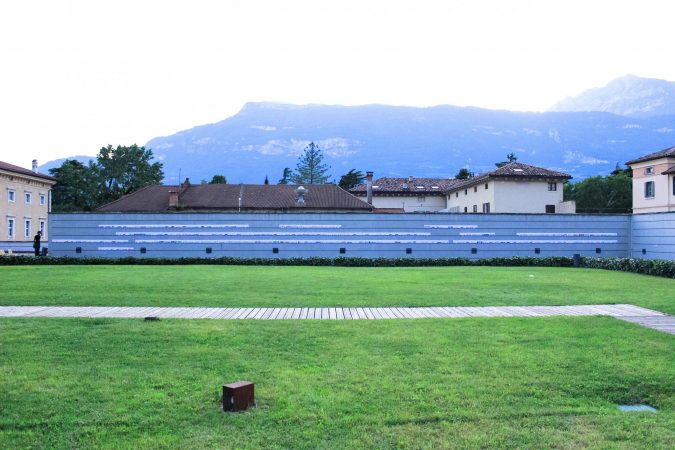A long gray concrete wall, in front of a grass field, is used to expose three long lines of paper sheets with drawings