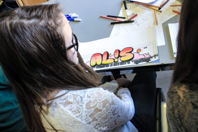 A young girl shows a graffiti drawing on the side of a train wagon, with her nickname drawn by herself on a paper sheet