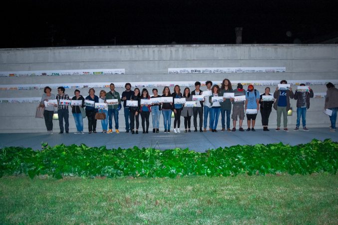 A big group of kids standing against a gray concrete wall each one of them holding a paper sheet in front of him to show his own drawing