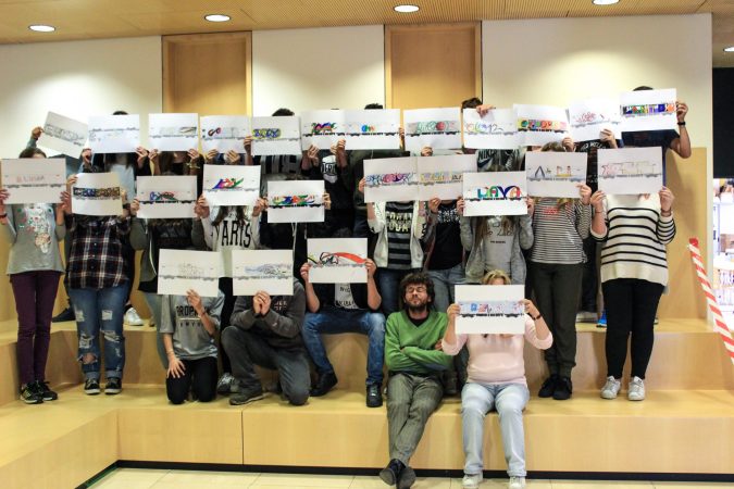A big group of kids sitting on benches disposed on three layers, every kid is displaying a paper sheet in front of his face to show his own drawing