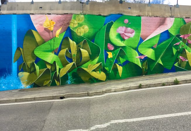 Total view of a Mural Painting, across a street, depicting the name of the city Covignano woven with colored flowers and green leaves. Artwork by Dado
