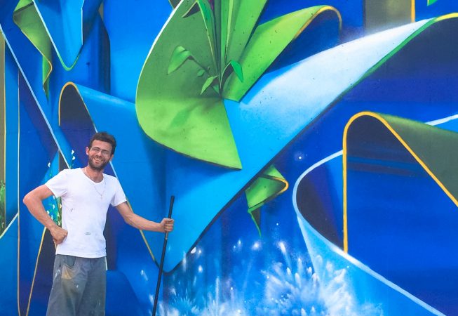 Dado Ferri posing in front of his freshly finished mural artwork depicting wide blue ribbons decorated with purple flowers and green leaves.