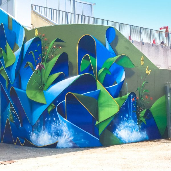 Total view of a large mural painting depicting wide sinuous blue ribbons decorated with purple, red and yellow flowers and green leaves and water splashes. Artwork by Dado