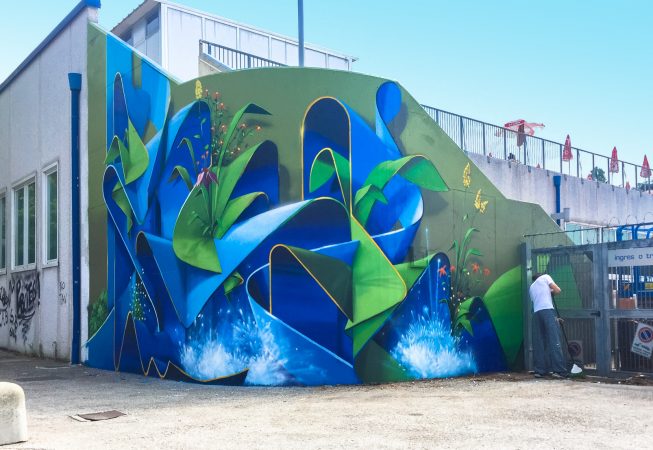 Wide location view of a large mural painting depicting wide sinuous blue ribbons decorated with purple, red and yellow flowers and green leaves and water splashes. Artwork by Dado