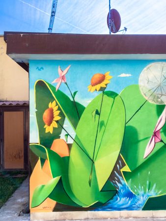 Detail of a mural painting depicting big green leaves with geometric rounded shapes and small pink and yellow flowers and a white spherical dandelion seeding head floating on deep blue water. Artwork by Dado