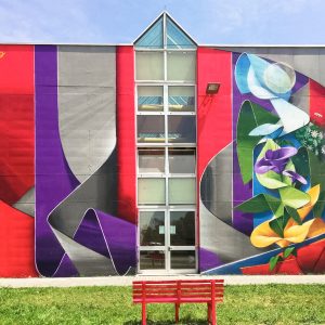 Front view of an high school building face with a mural painting depicting wide gray and purple ribbons composing the name Piero in a graffiti wildstyle decorated with yellow and purple flowers. Artwork by Dado.