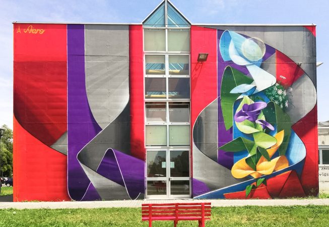 Front view of an high school building face with a mural painting depicting wide gray and purple ribbons composing the name Piero in a graffiti wildstyle decorated with yellow and purple flowers. Artwork by Dado.