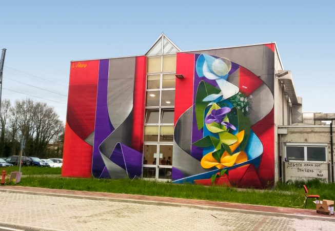 Lateral view of an high school building face with a mural painting depicting wide gray and purple ribbons composing the name Piero in a graffiti wildstyle decorated with yellow and purple flowers. Artwork by Dado.