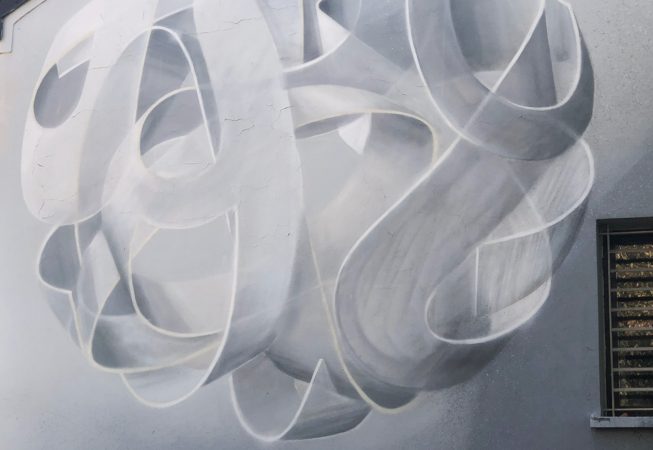 Main element of a mural painting depicting a spherical wildstyle graffiti forming the tag Dado colored in a very vanishing white and light gray tones.