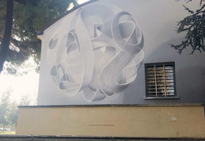 Total view of a mural painting depicting a spherical wildstyle graffiti forming the tag Dado colored in a very vanishing white and light gray tones.