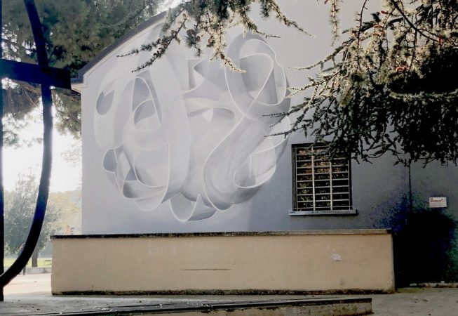 A wall behind trees with a mural painting depicting a spherical wildstyle graffiti forming the tag Dado colored in a very vanishing white and light gray tones.