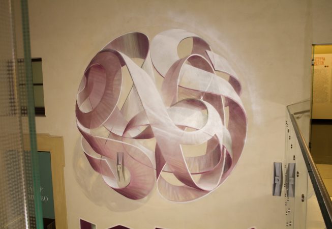 Mural painting inside a depicting a spherical wildstyle graffiti forming the tag Dado colored in a vanishing light pink and winy tones.