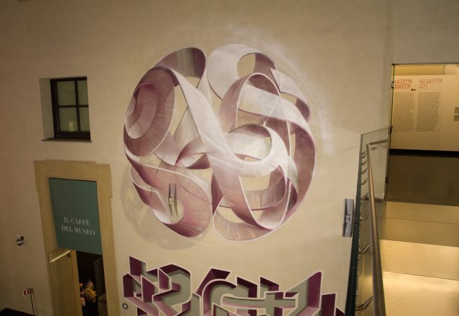 Total top view of a mural painting inside an art gallery depicting a spherical wildstyle graffiti forming the tag Dado colored in a vanishing light pink and winy tones.