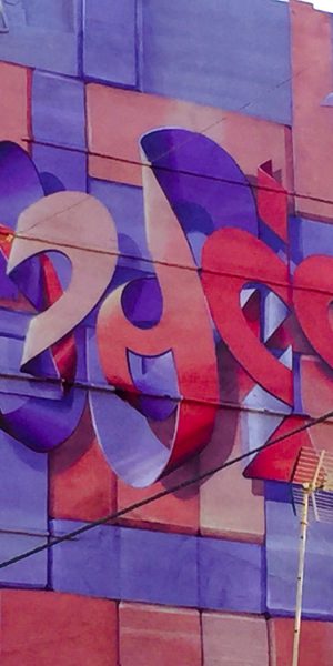 Bottom view of a Mural painting on an high building face depicting floating bent ribbons creating two names altogether the tag Dado and the name Eva in a pale red and electric blue tones