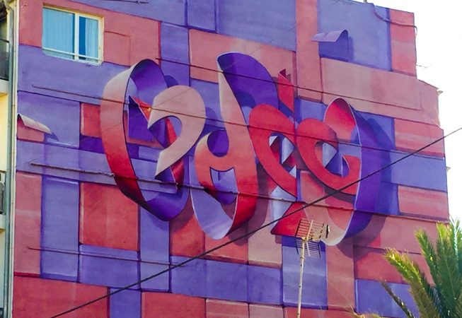 Bottom view of a Mural painting on an high building face depicting floating bent ribbons creating two names altogether the tag Dado and the name Eva in a pale red and electric blue tones