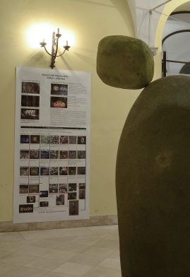 Detail of small yellow stone on top of another bigger stone, positioned in an impossible balance close to fall down the floor. In the background is visible a poster, stitched to a wall, where is explained the "equation" that stands behind the artwork. Sculpture by Dado