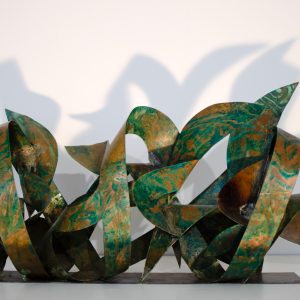 Front view of a sculpture made of copper ribbons sinuously coiled, forming the tag of the artist Dado. The copper colored surface is texturized with extensive green oxidation stains.