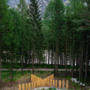 Panoramic view of a mountain landscape with a series of tall wooden boards planted in the ground forming a big X in the middle of a mountain path, in front of a grass field, with high trees in the background. Sculpture installation by Dado.